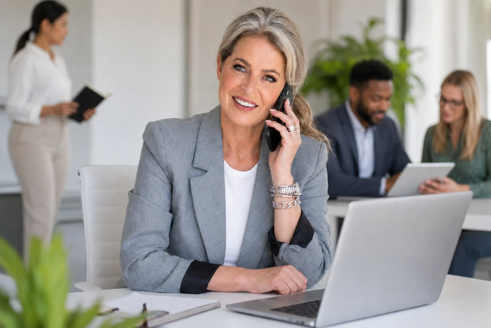 Een ervaren professional spreekt een klant telefonisch en werkt achter een laptop, met collega’s op de achtergrond.